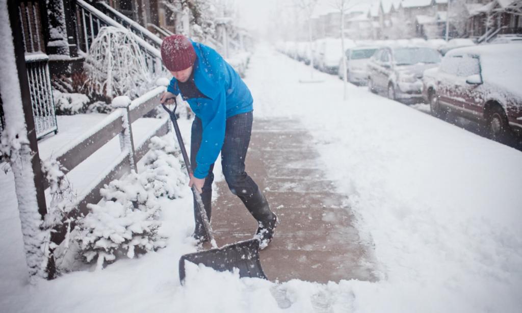 Мужчина с синей куртке чистит снег лопатой